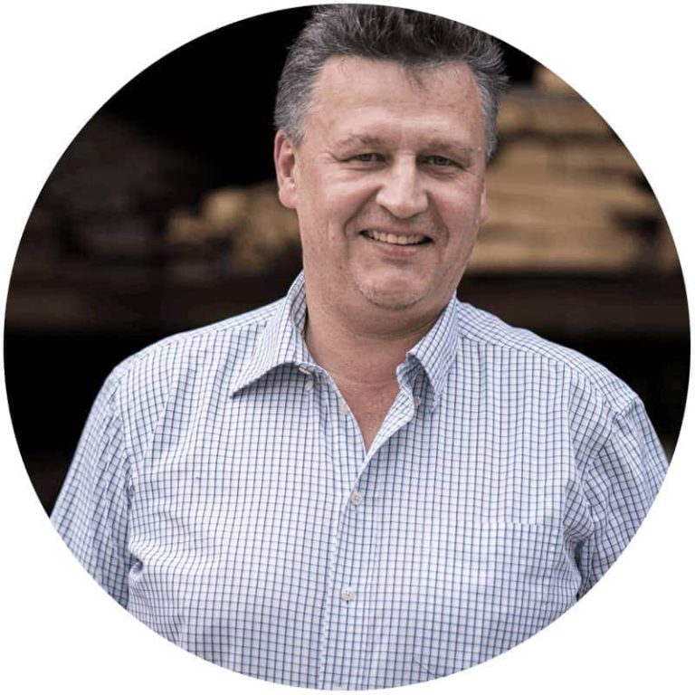 Lächelnder Mann mit kariertem Hemd vor Holzwerkstatt, repräsentiert Holzbau Bachmaier.